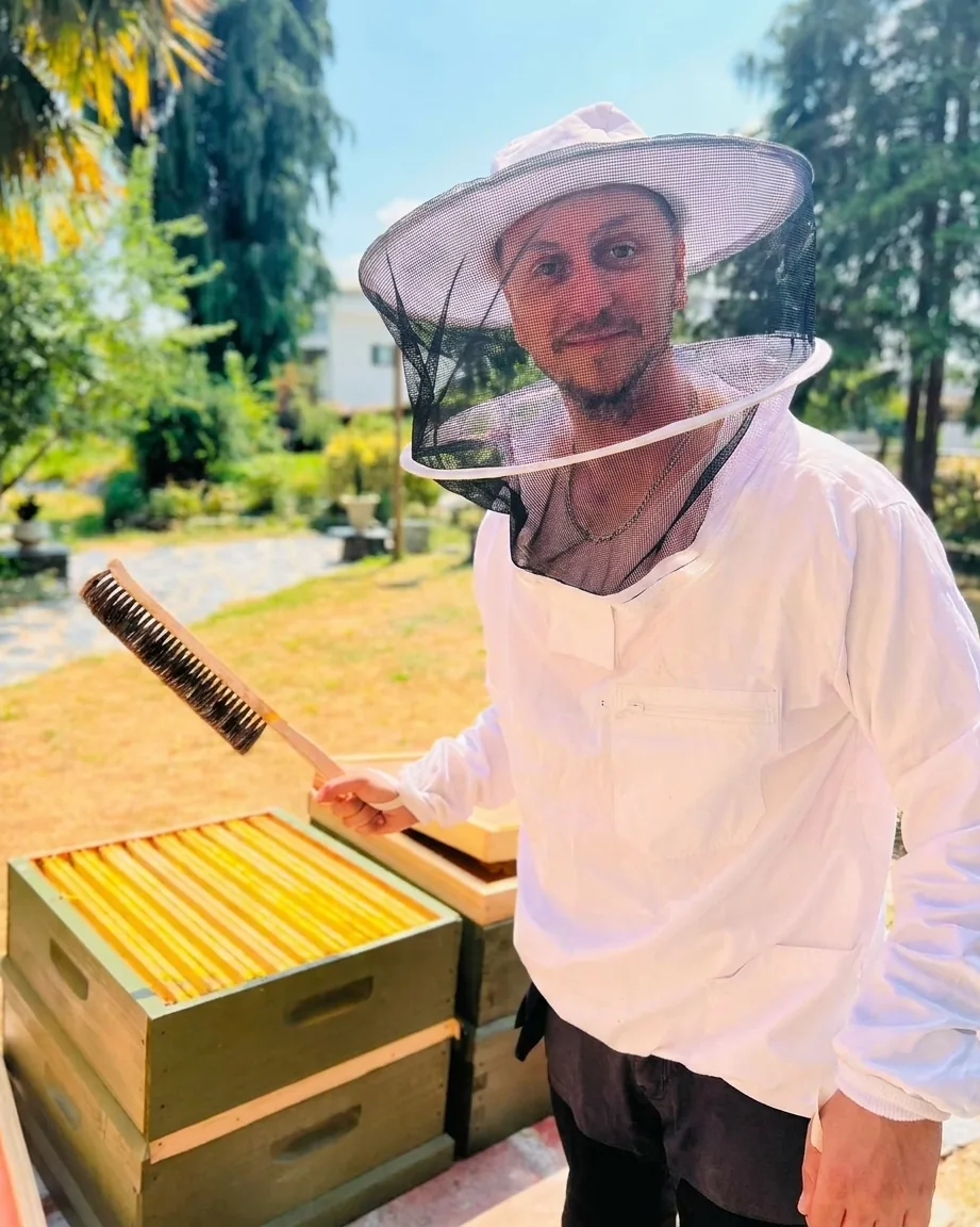 L’apiculteur d’AS MIEL en vareuse, brosse à abeilles en main, devant une ruche ouverte du jardin